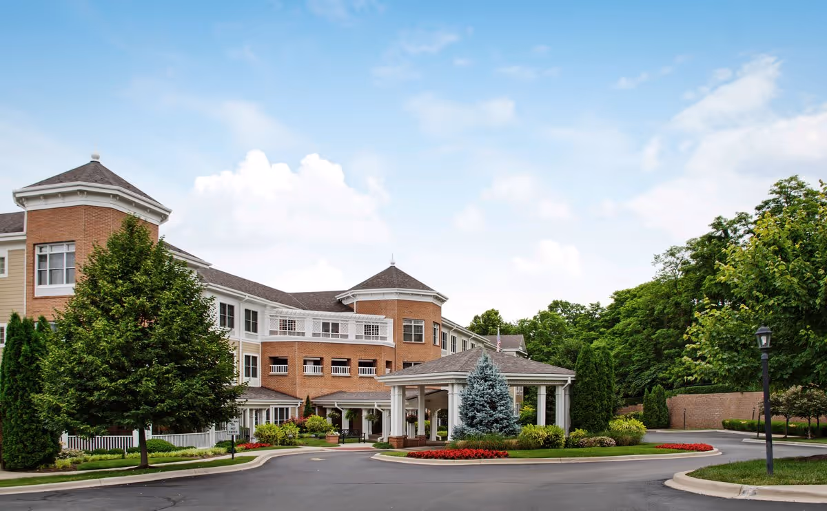 Exterior front entrance of a multi-story senior living building with a circular drive, covered porte-cochère, and landscaped trees and shrubs.