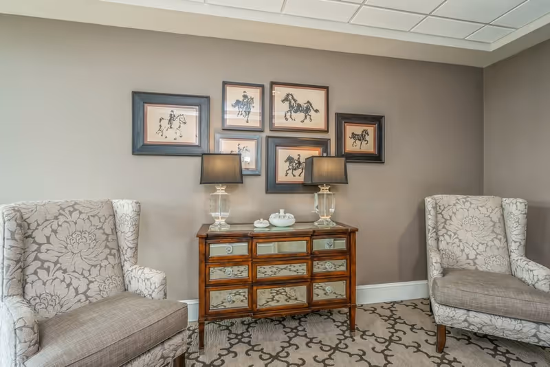 A cozy sitting area with two patterned armchairs facing each other, a wooden mirrored dresser with two table lamps on top, and six framed pictures of horses on the wall above the dresser. The room has a beige wall and a patterned carpet.