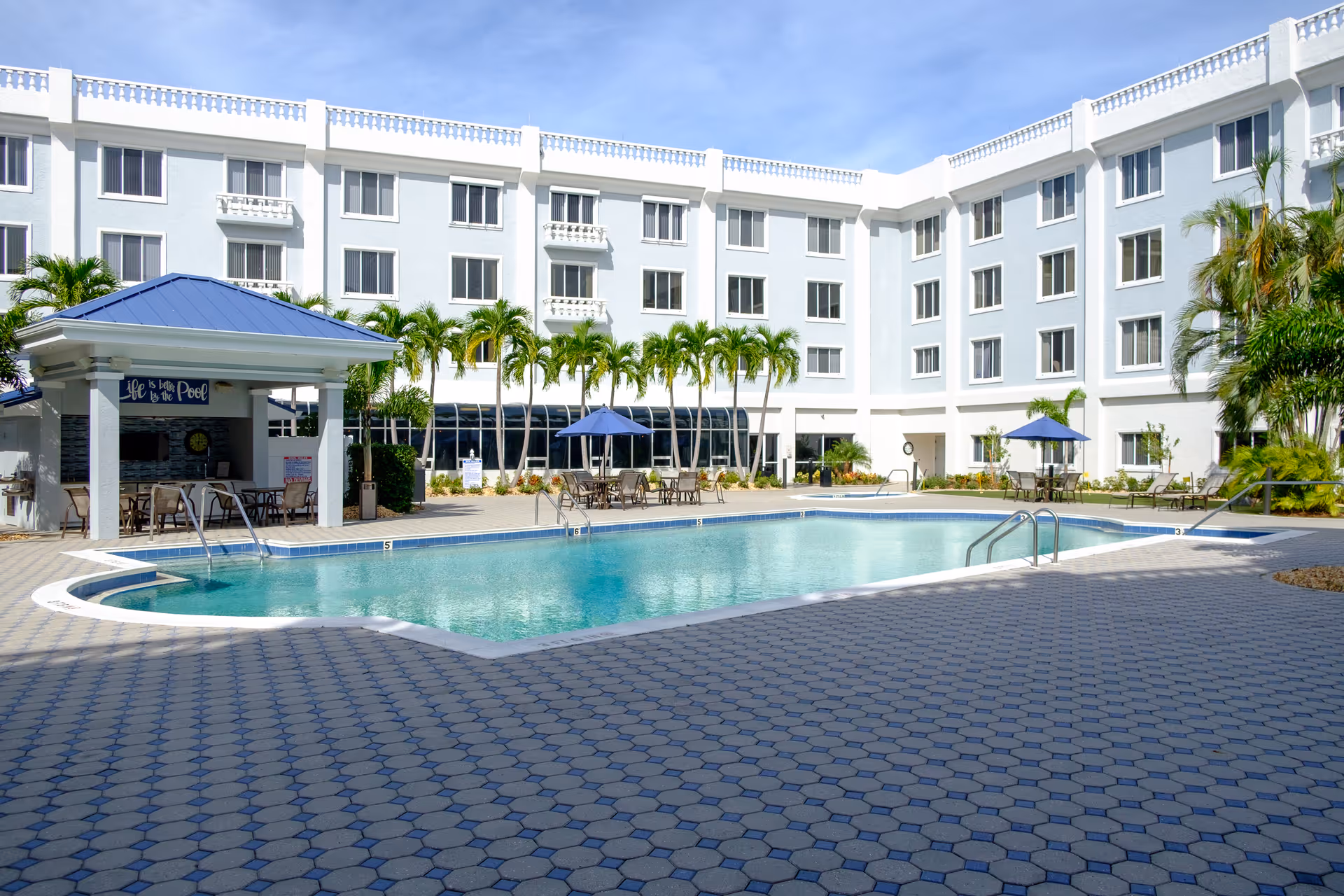 Outdoor swimming pool area at The Summit of Fort Myers with a covered seating area, tables with umbrellas, palm trees, and a multi-story white building in the background under a clear blue sky.