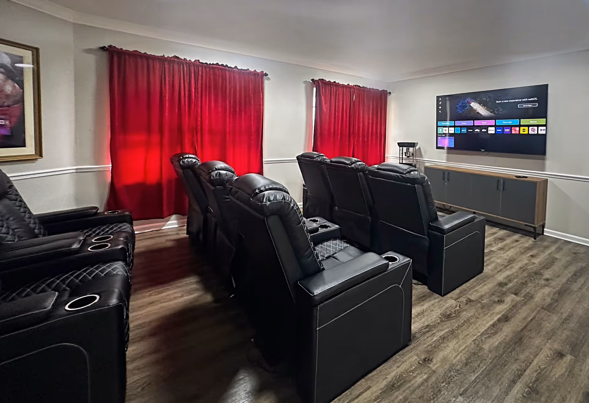 A cozy media room with two rows of black leather recliner chairs facing a wall-mounted flat-screen TV. The room has wood flooring, red curtains covering the windows, and a modern console table beneath the TV.