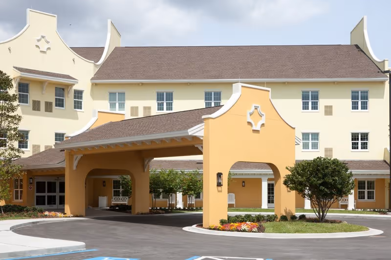 Exterior view of Discovery Village Trinity senior living facility showing a large covered entrance with yellow and beige walls, multiple windows, landscaped flower beds, and a tree near the driveway.