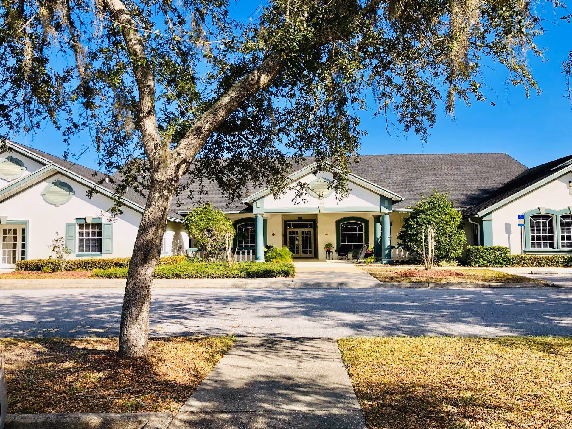 Front exterior view of a single-story building with a peaked roof and green trim around windows and doors. There is a large tree in the foreground with branches extending over the sidewalk leading to the entrance. The entrance has a covered porch supported by green columns, and there are shrubs and small trees around the building. The sky is clear and blue.