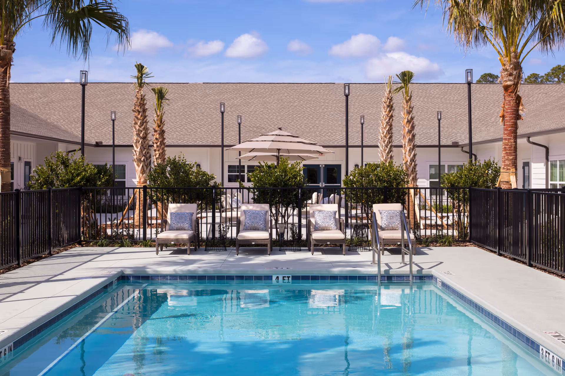 Outdoor swimming pool area with clear blue water, surrounded by a black metal fence. Four cushioned lounge chairs with patterned pillows are lined up on the pool deck. Behind the chairs, there are palm trees, green bushes, and a patio area with an umbrella and additional seating. The building in the background has a light-colored exterior and a sloped roof under a partly cloudy sky.