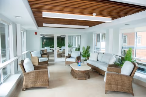 A bright and airy indoor seating area with wicker furniture including a sofa, four armchairs, and a central coffee table. The room features large windows on two sides, letting in natural light, and a wooden slatted ceiling with modern lighting fixtures. There are green plants placed near the windows and glass doors leading to another room with similar seating.
