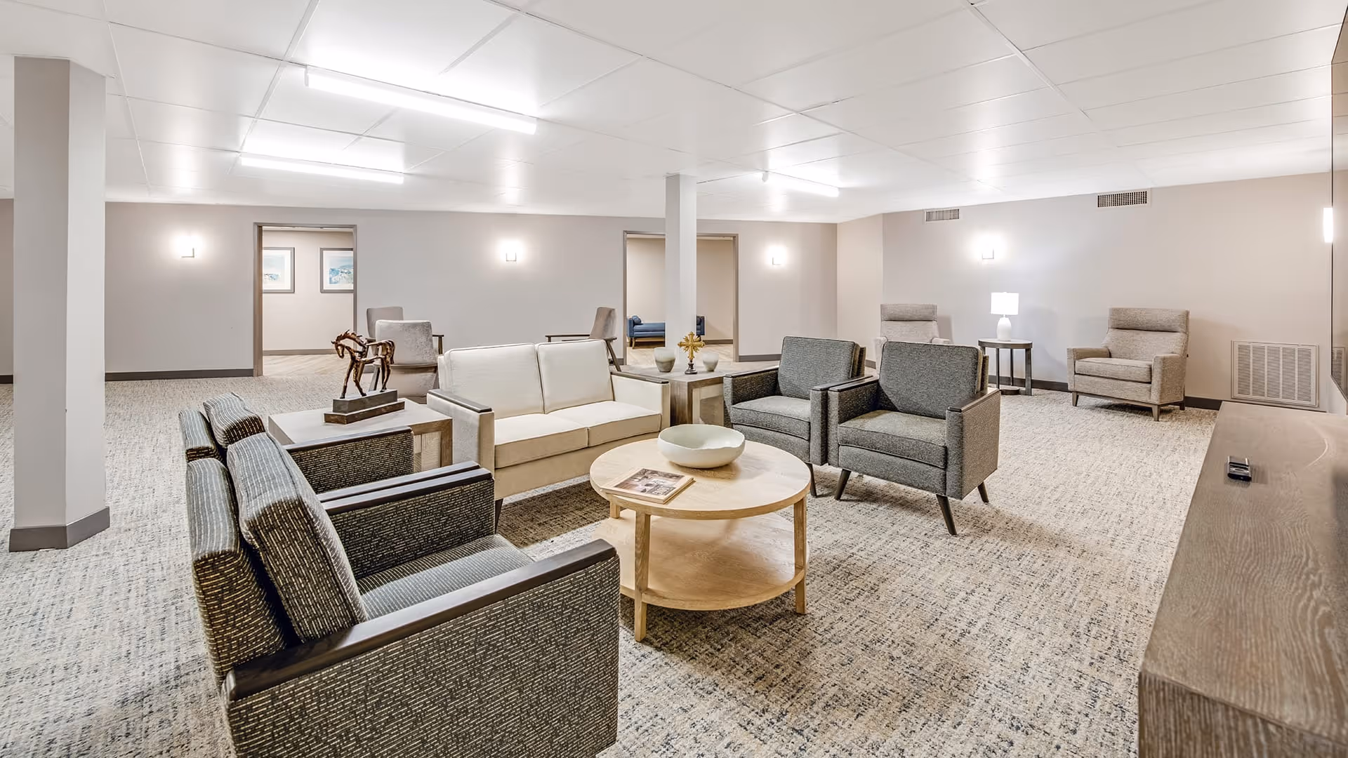 A spacious and well-lit living room area in a senior living facility with multiple armchairs and sofas arranged around a round wooden coffee table. The room has light-colored walls, carpeted floors, and several wall-mounted lights. There are doorways leading to other rooms in the background.