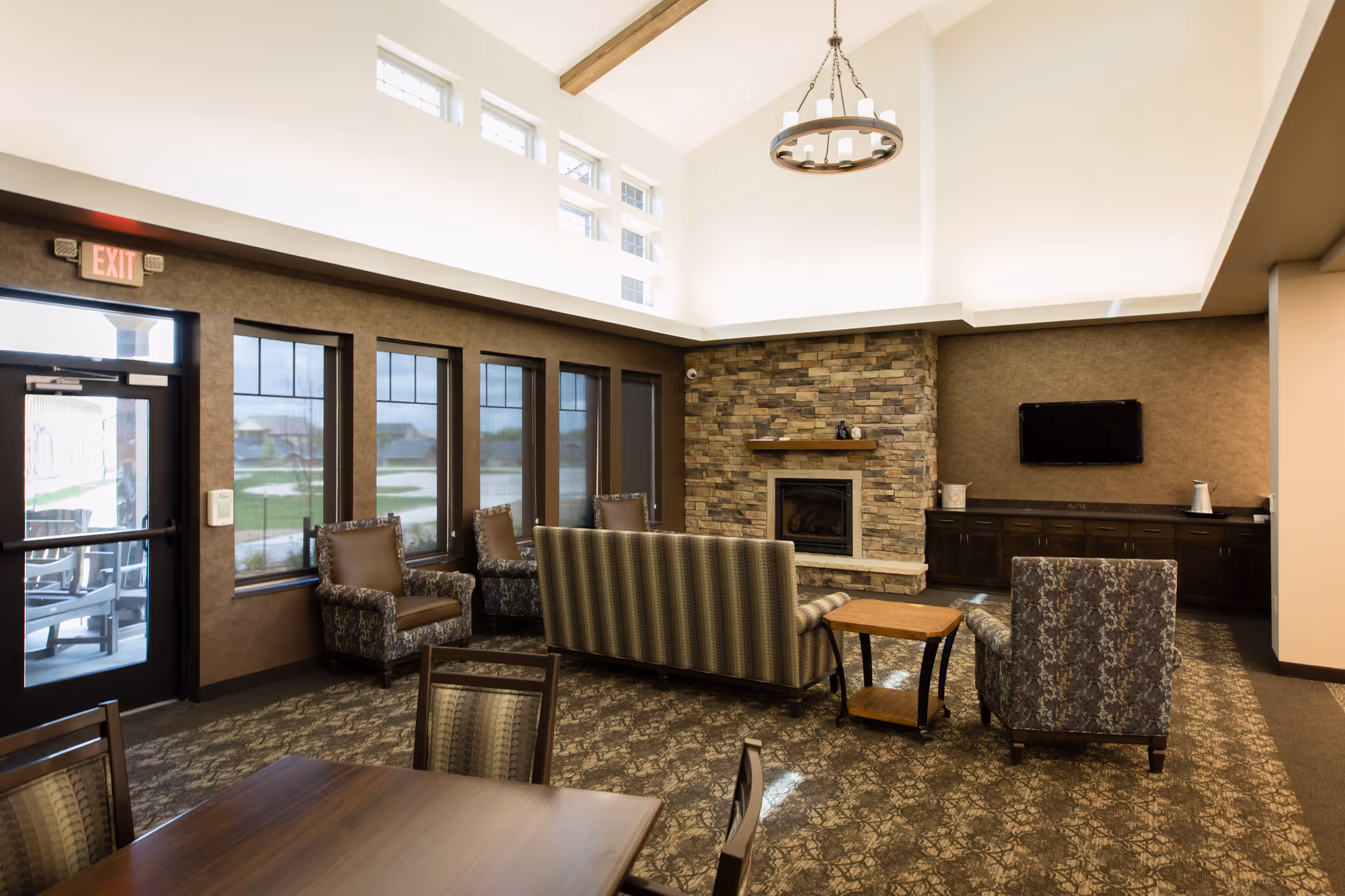 A cozy living room area in The Ranch House facility featuring a stone fireplace, a mounted flat-screen TV, several upholstered chairs and a sofa arranged around a wooden side table, large windows with shades, and a high ceiling with a circular chandelier.