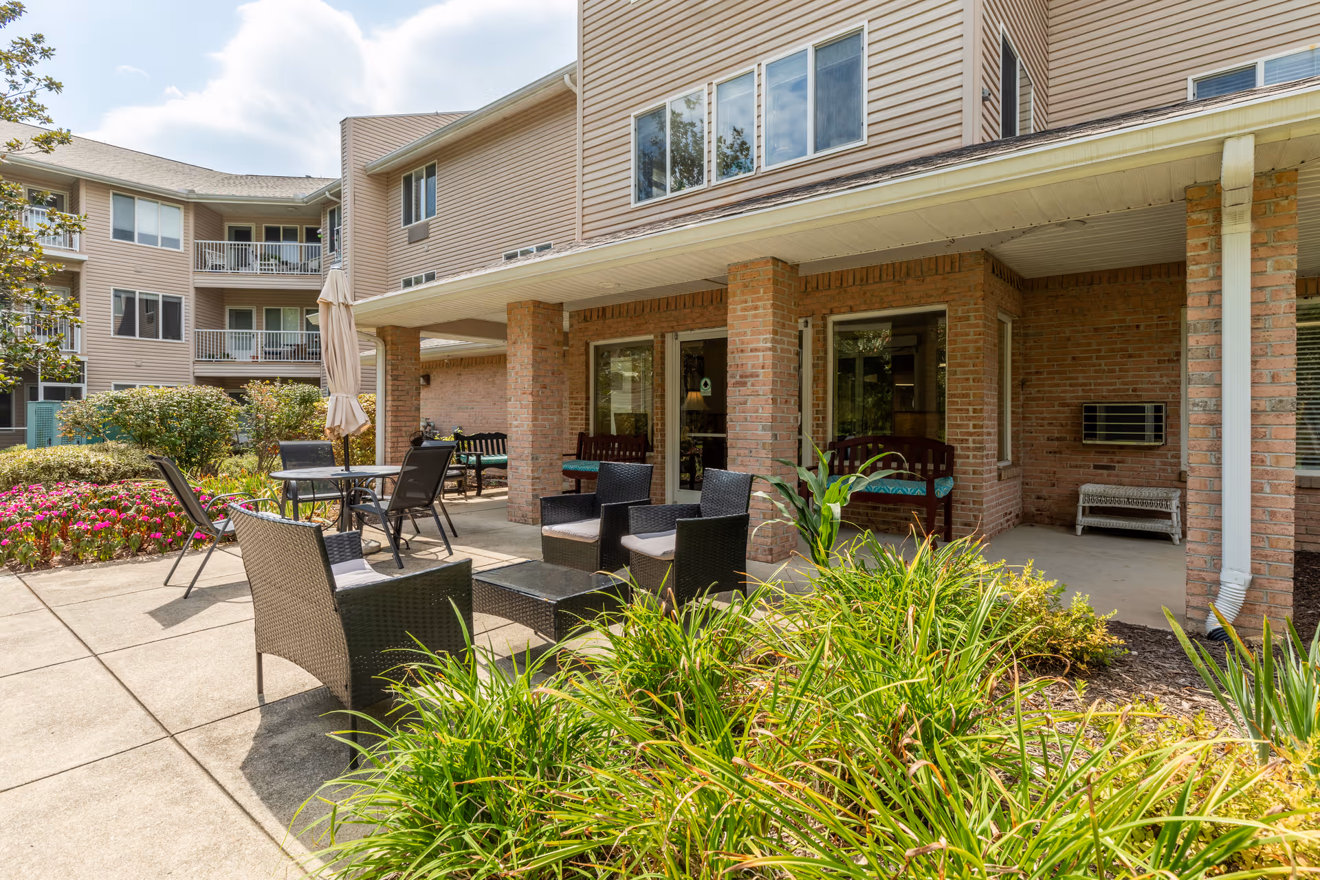 Outdoor patio area at a senior living community with several black wicker chairs and tables, some with cushions, surrounded by green plants and flowers. The patio is adjacent to a brick building with large windows and a covered porch supported by brick columns.