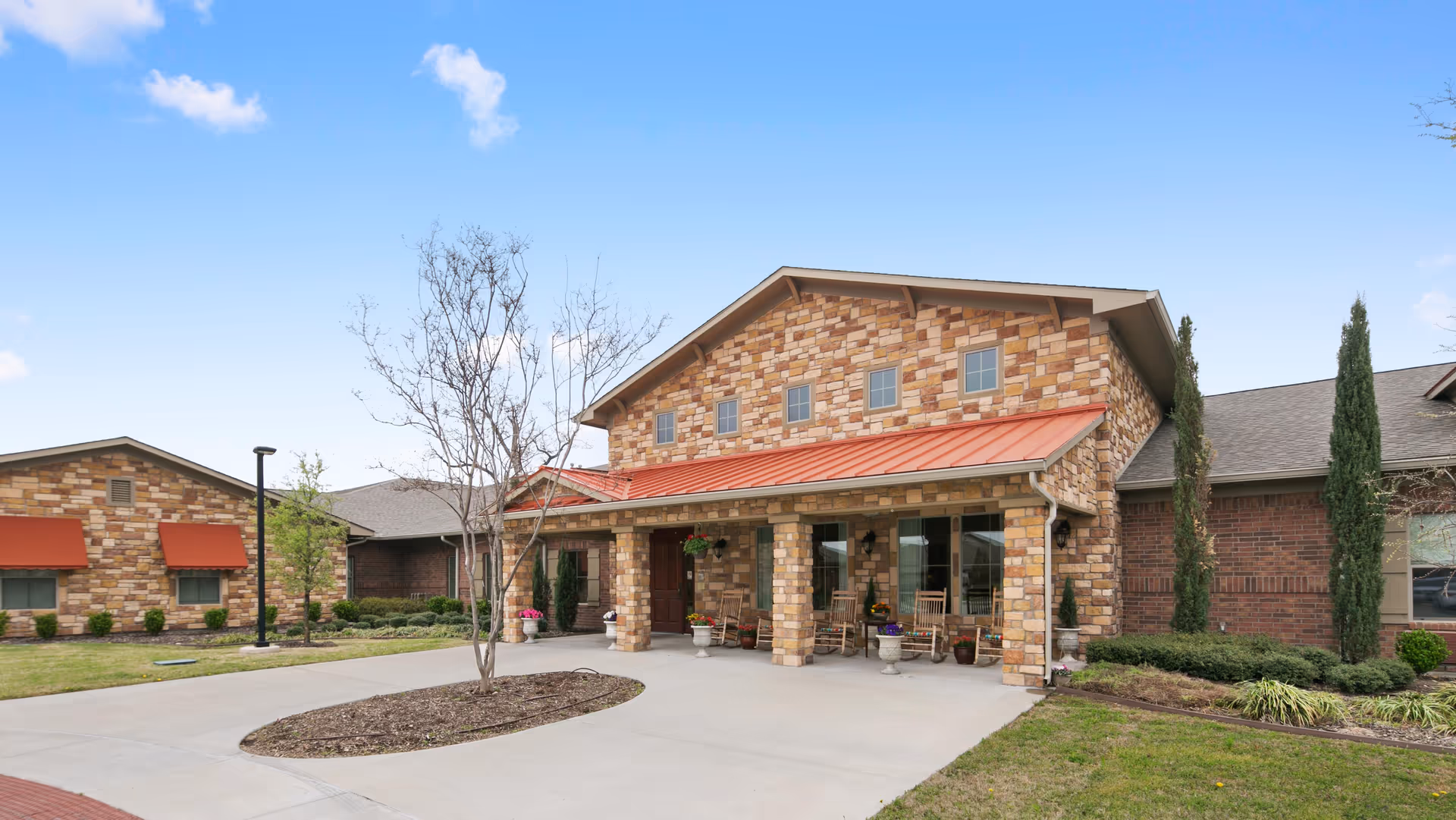 Stone-front assisted living building entrance with a covered porch, rocking chairs, driveway and landscaped grounds.