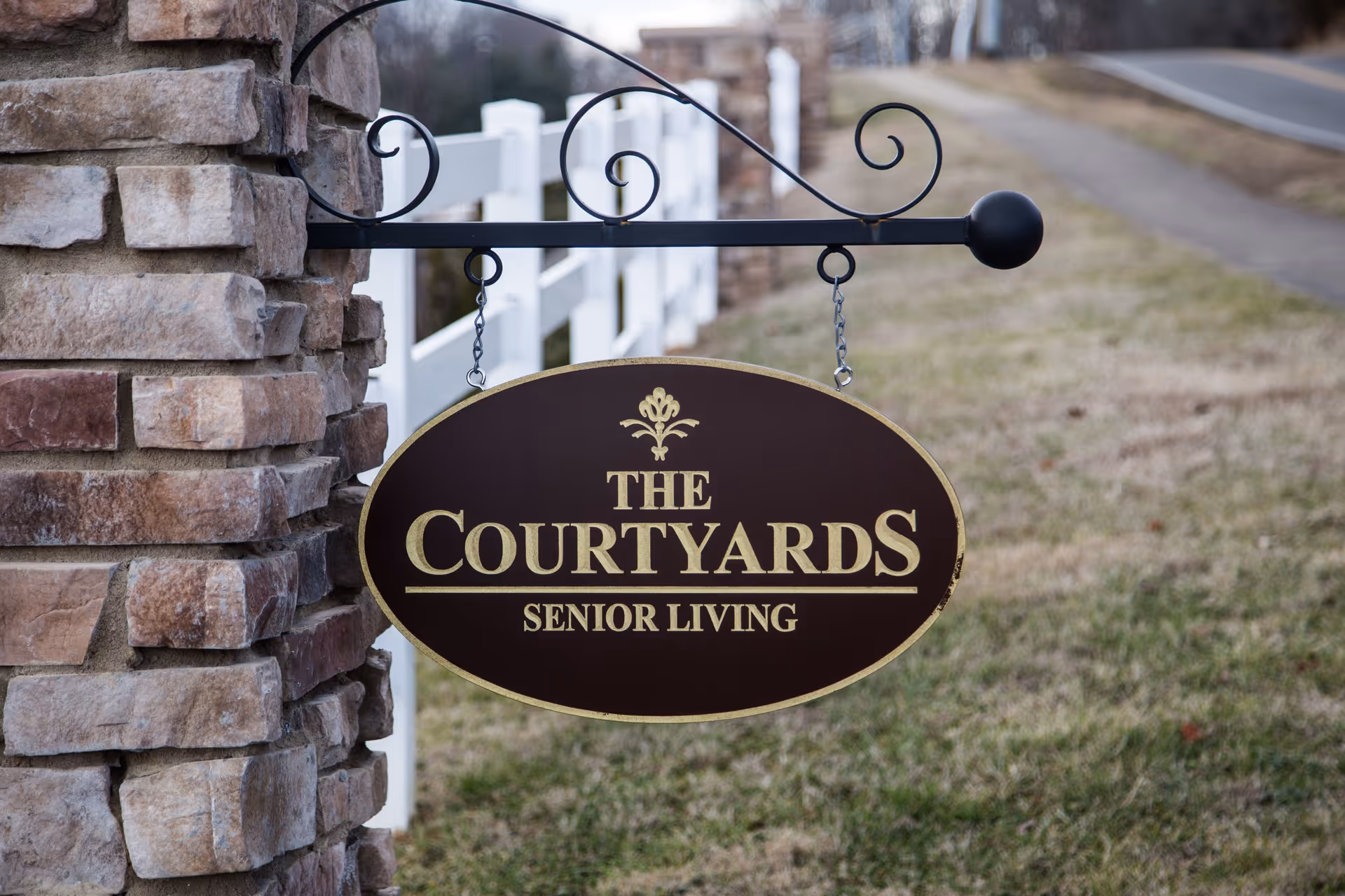 A hanging oval sign attached to a stone pillar with the text 'The Courtyards Senior Living' in gold letters, with a white fence and grassy area in the background.