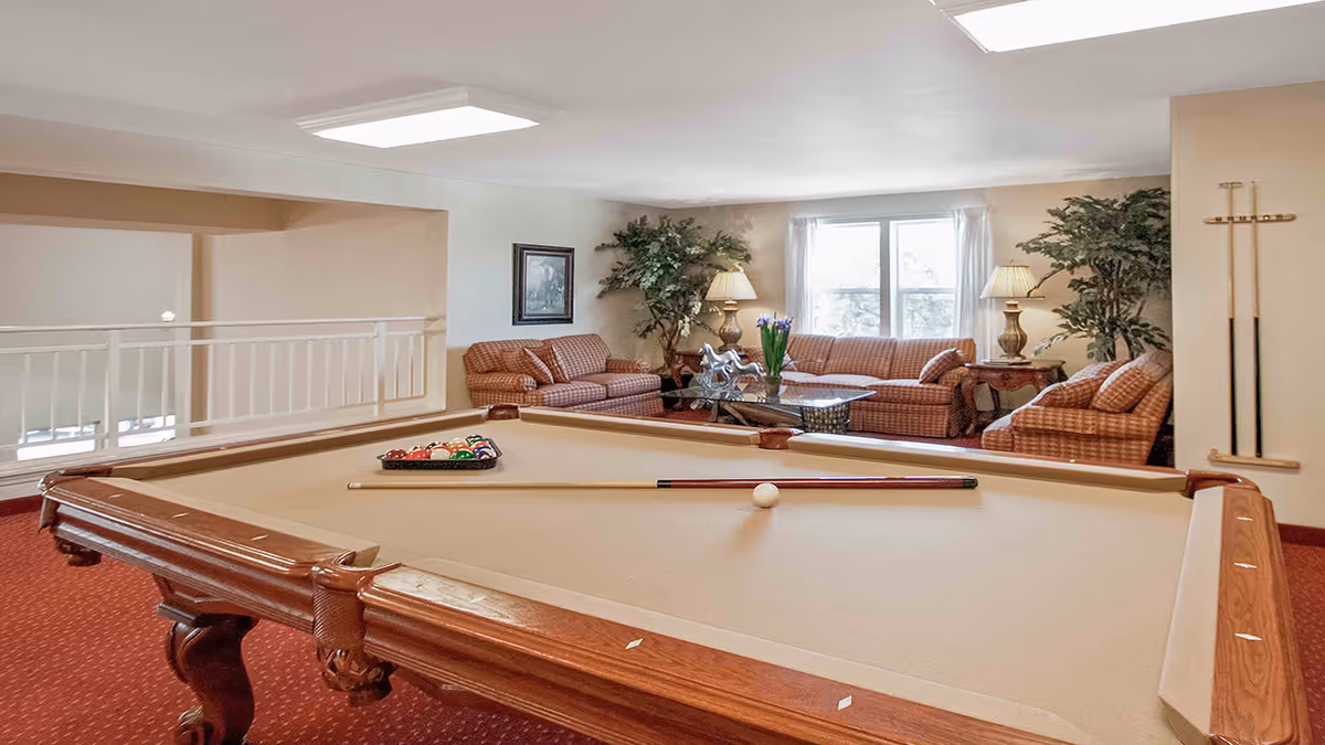 A pool table in the foreground with a comfortable lounge area of sofas, lamps and plants by a window in the background.