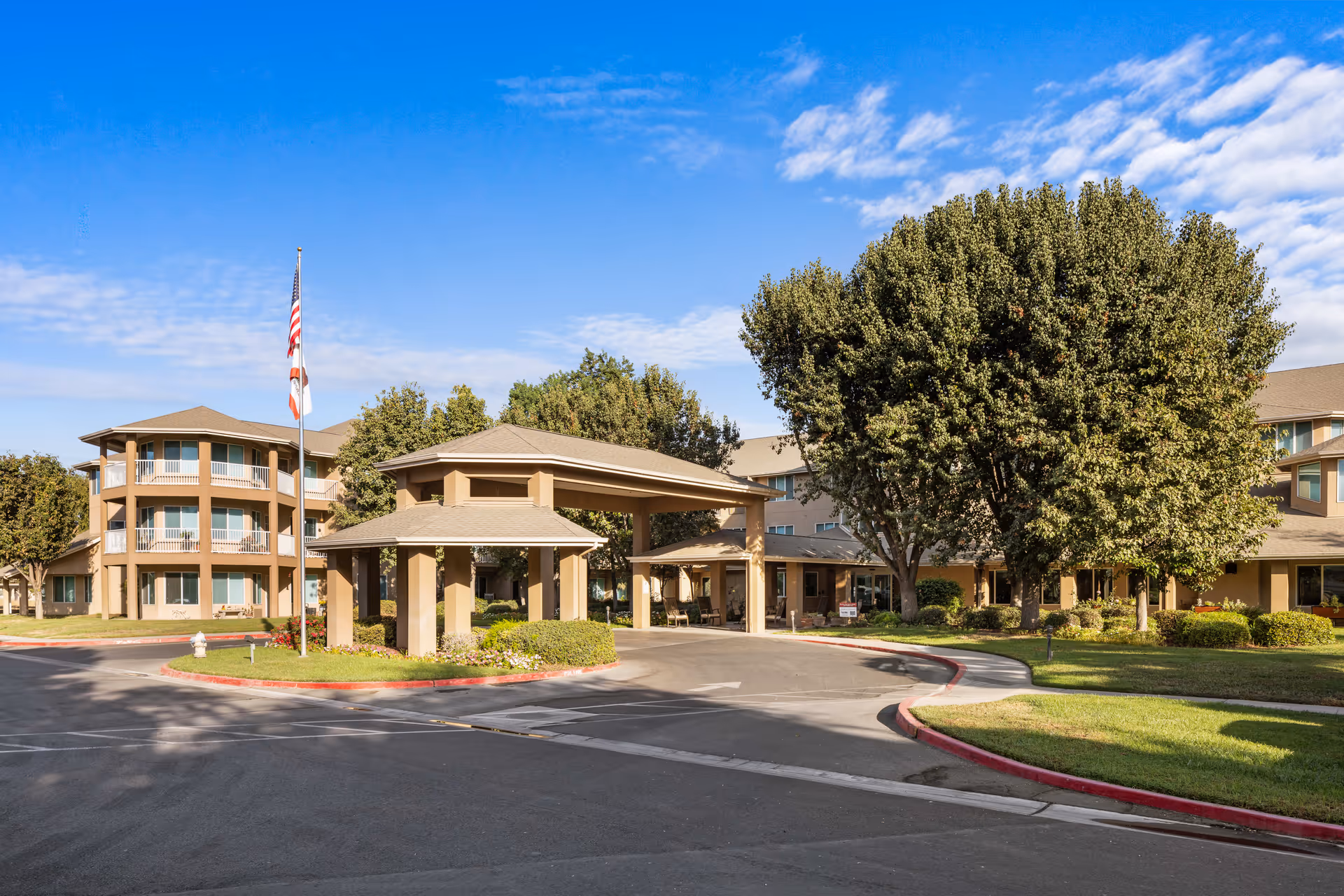 Exterior view of Sierra Hills - A Provincial Senior Living Community, showing a multi-story building with balconies, a covered entrance driveway, well-maintained landscaping with trees and bushes, and an American flag on a flagpole under a blue sky with some clouds.
