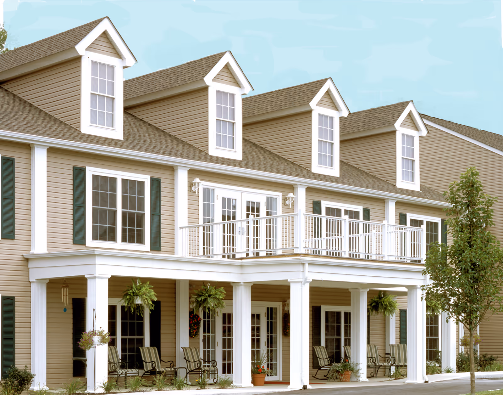 Exterior view of a beige two-story building with white trim and green shutters. The building features a covered porch with several chairs and hanging plants, and a balcony with white railings on the second floor. There is a small tree and some landscaping in front of the building under a clear blue sky.