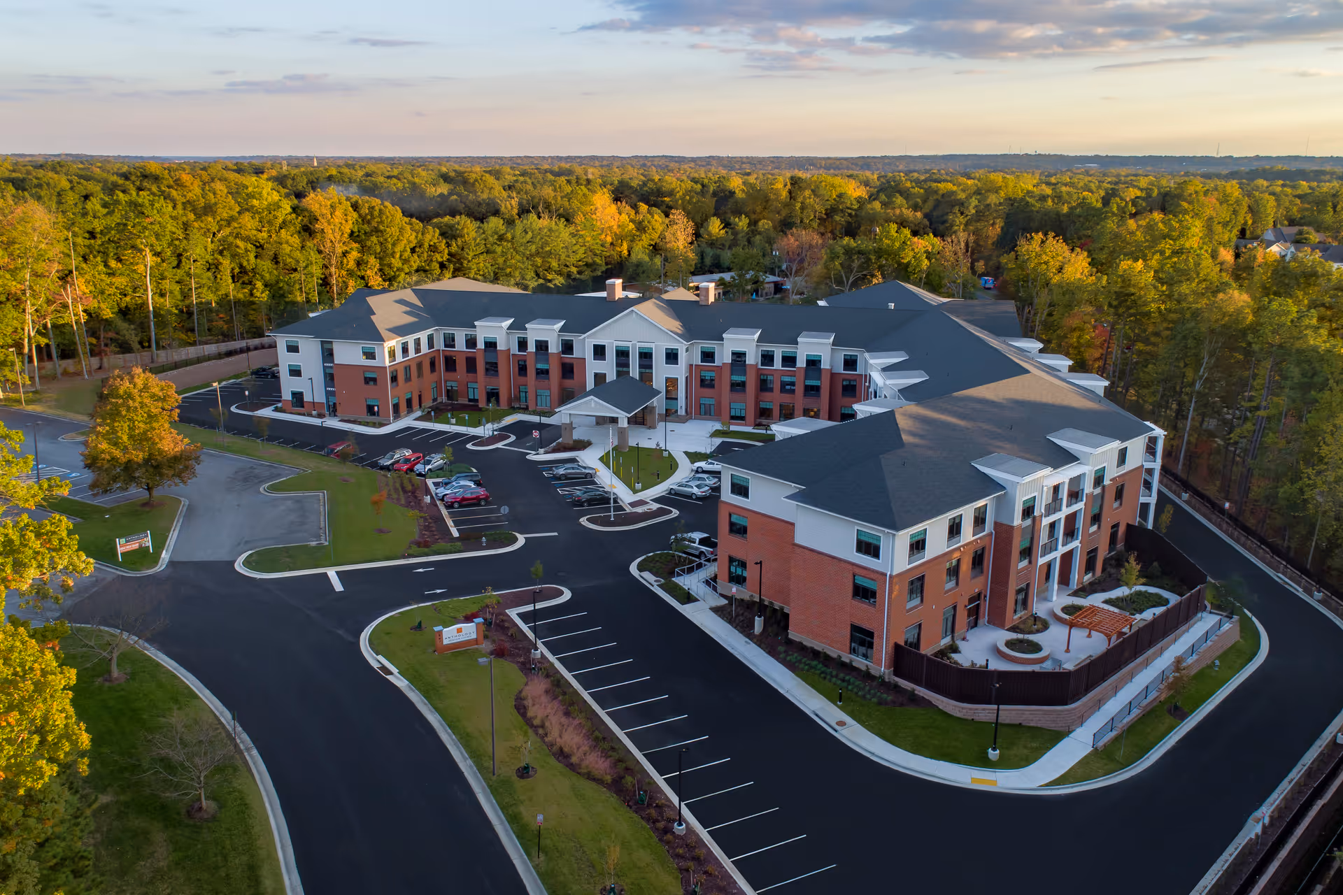 Aerial view of Elancé at Tuckahoe, a large senior living facility surrounded by trees. The building is three stories tall with a combination of red brick and white siding exterior. There is a parking lot with several cars and landscaped areas around the building. The sky is partly cloudy with soft evening light.