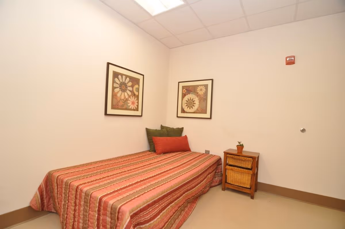 A small bedroom with a single bed covered in a striped red, pink, and brown bedspread. There are three pillows on the bed, two green and one red. On the wall above the bed are two framed floral artworks. Next to the bed is a small wooden nightstand with two wicker baskets and a small potted plant on top. The walls are plain white, and the ceiling has a grid pattern with fluorescent lighting.