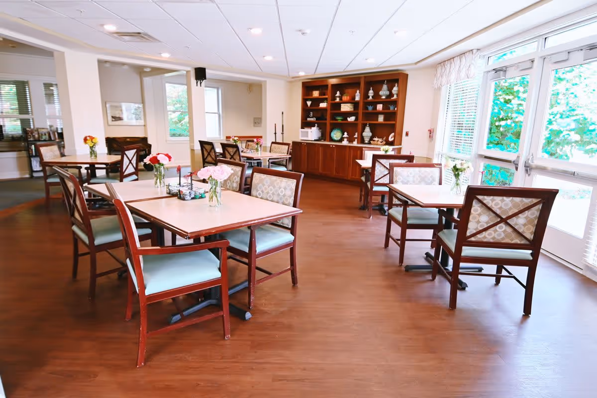 A bright dining room featuring several wooden tables with chairs, each adorned with small flower vases. A wooden shelf in the background displays decorative items, and large windows allow natural light to illuminate the space.