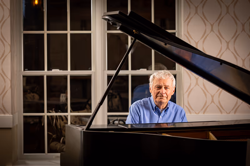An elderly man with white hair wearing a blue shirt is sitting at a grand piano in a room with patterned wallpaper and large windows behind him.