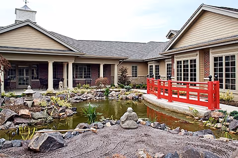 Outdoor courtyard area of a senior living facility featuring a pond with rocks and plants, a small stone statue, and a red wooden bridge. The building surrounding the courtyard has beige siding with brick accents and large windows.