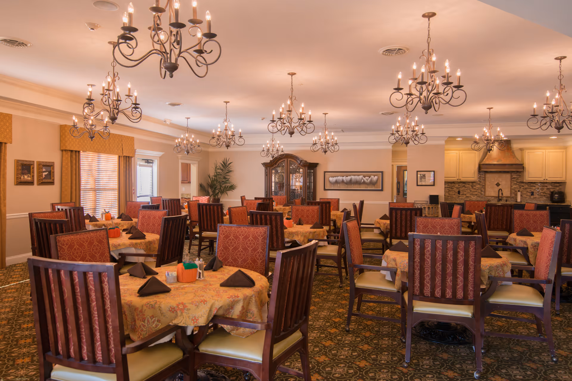 Spacious, warmly lit dining room with multiple round tables, patterned chairs, and ornate chandeliers.