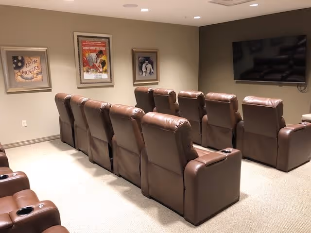 A small theater room with two rows of brown leather recliner chairs facing a large flat-screen TV mounted on the wall. The walls are beige and decorated with three framed vintage movie posters.