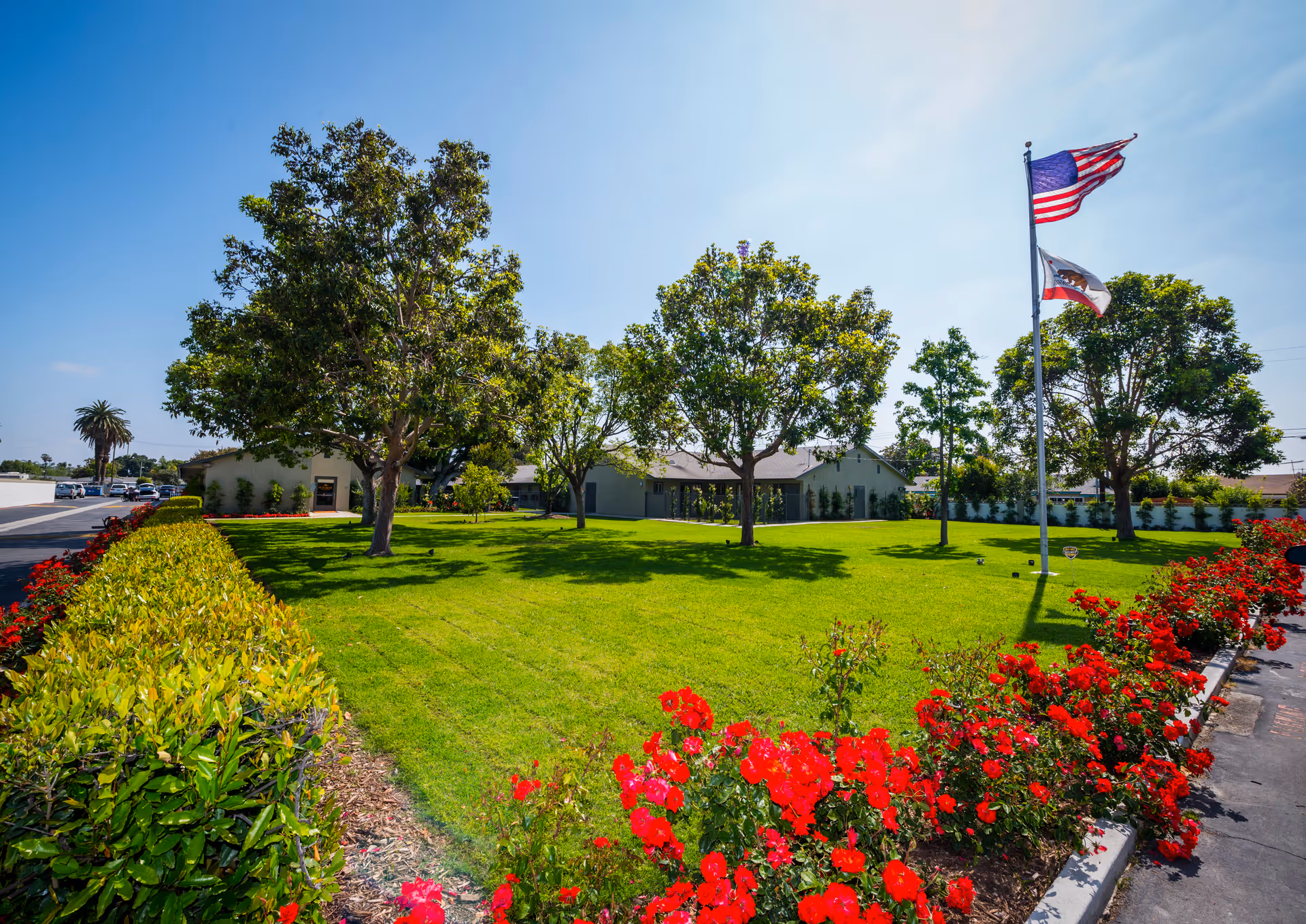 A well-maintained outdoor garden area with lush green grass, several trees, and vibrant red flowers along the edge. Two flagpoles display the American flag and the California state flag. A building is visible in the background under a clear blue sky.