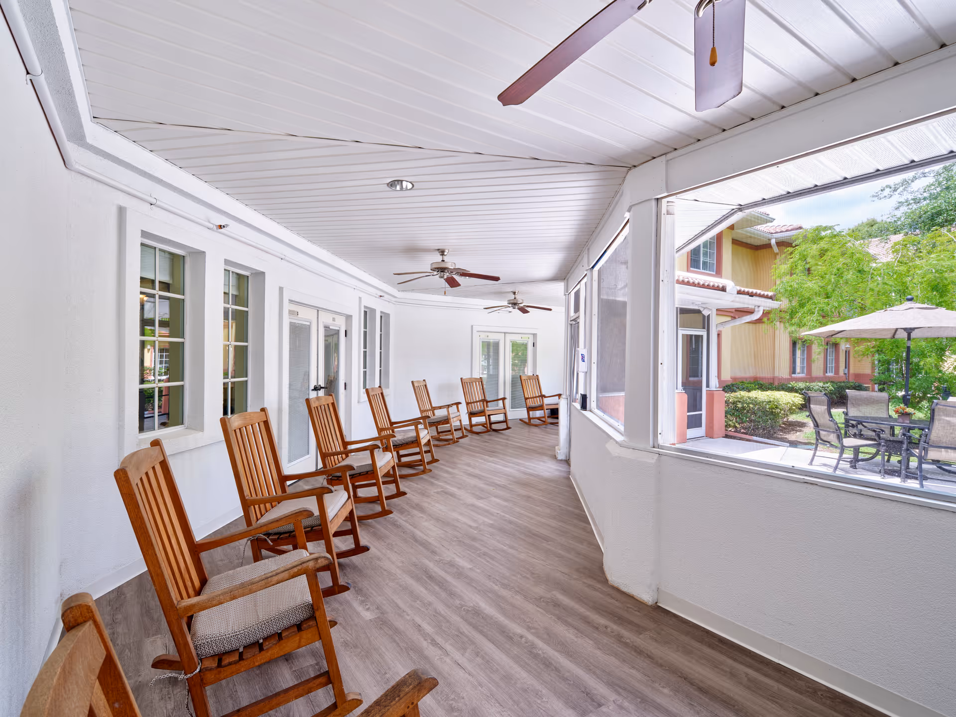 A covered porch area with wooden rocking chairs lined up along a white wall with windows and doors. The porch has a wood-like floor, ceiling fans, and large screened windows looking out to an outdoor patio with tables, chairs, and umbrellas surrounded by greenery and a building with a tiled roof.