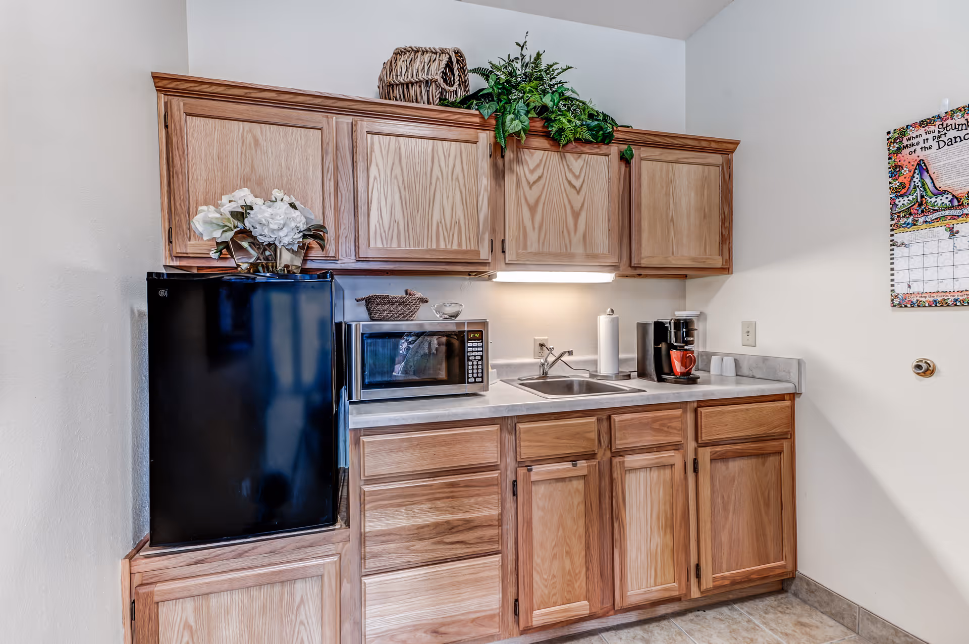 A small kitchen area with wooden cabinets, a black mini refrigerator, a microwave, a sink, a coffee maker with a red mug, and decorative plants on top of the cabinets. There is a colorful calendar on the wall to the right.