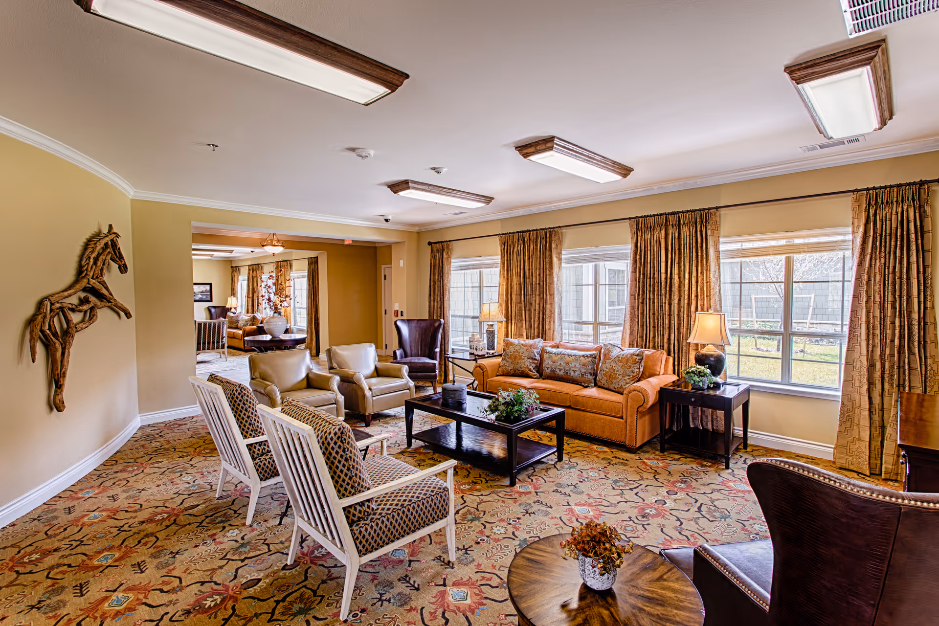 A spacious and well-lit living room in an assisted living facility featuring a large orange sofa with patterned cushions, two beige armchairs, two white wooden chairs with patterned cushions, a dark wooden coffee table with decorative plants, and large windows with brown curtains letting in natural light. The room has a patterned carpet and a wooden horse wall decoration on the left wall.