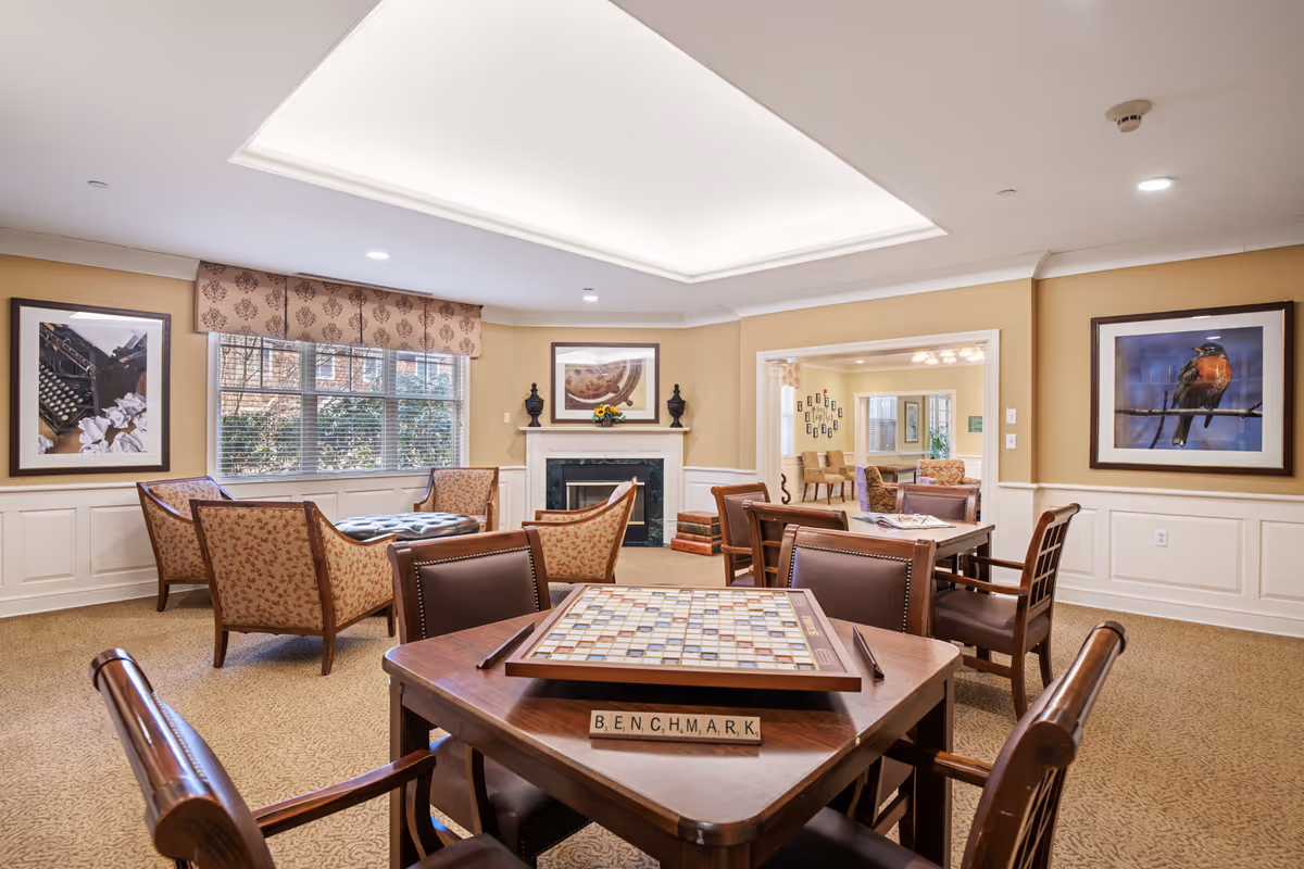 A cozy senior living common area with a ceiling light panel, beige walls, and carpeted floor. The room features several wooden tables with chairs, one table has a Scrabble board with letter tiles spelling 'BENCHMARK'. There are framed pictures on the walls, a window with patterned valance, and a fireplace with decorative items on the mantel. Another seating area is visible through an open doorway.