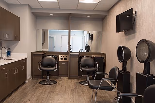 Interior of a salon area with three black salon chairs, two hair drying stations, a wall-mounted TV, a large mirror, and cabinetry with a sink on the left side.
