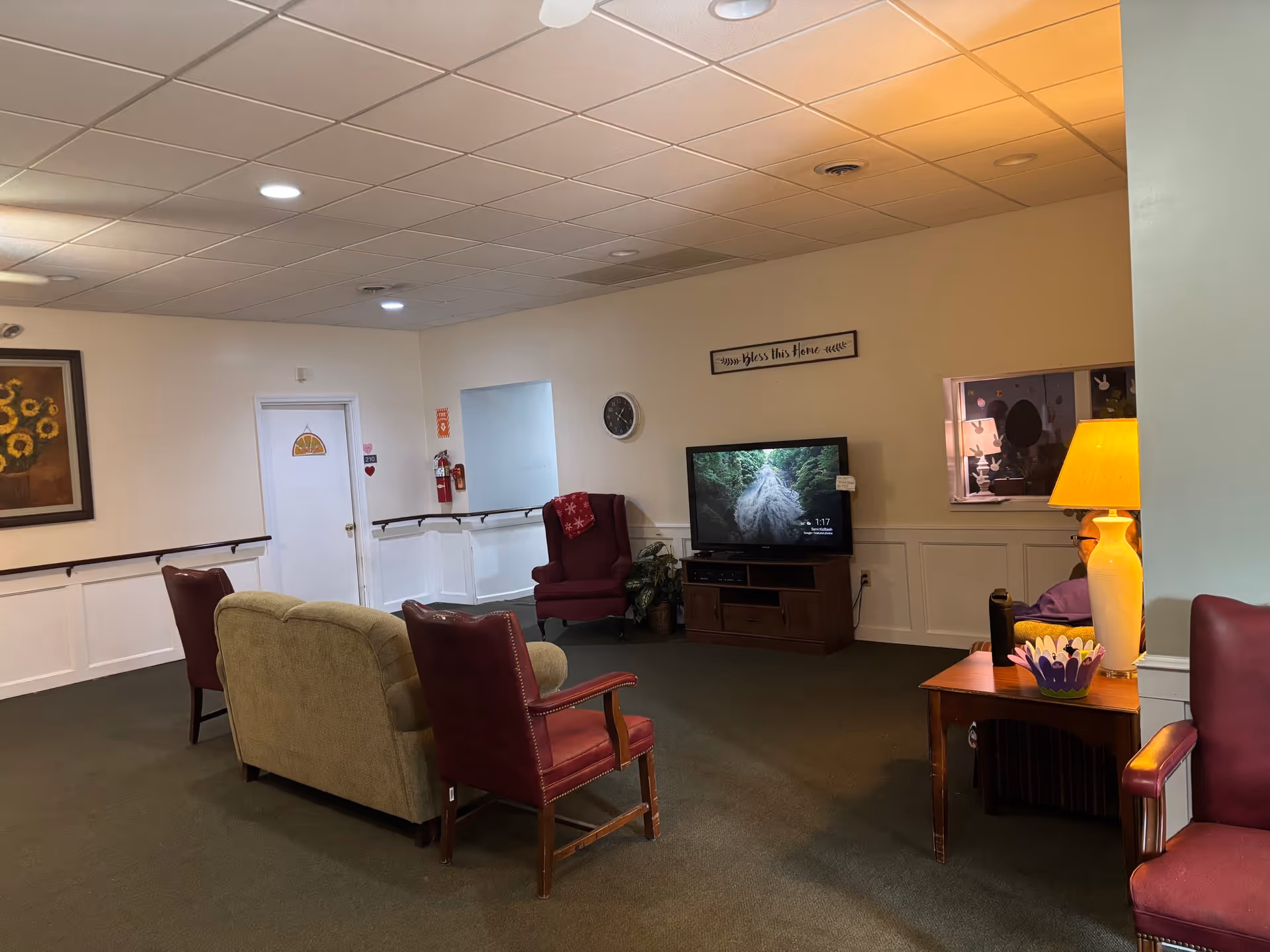 A senior living facility common area with a beige couch, two red armchairs, a wooden TV stand with a flat-screen TV displaying a nature scene, a wall clock, a lamp on a side table, and a sign on the wall that reads 'Bless this Home'. There is a painting of sunflowers on one wall and a fire extinguisher mounted near a white door.