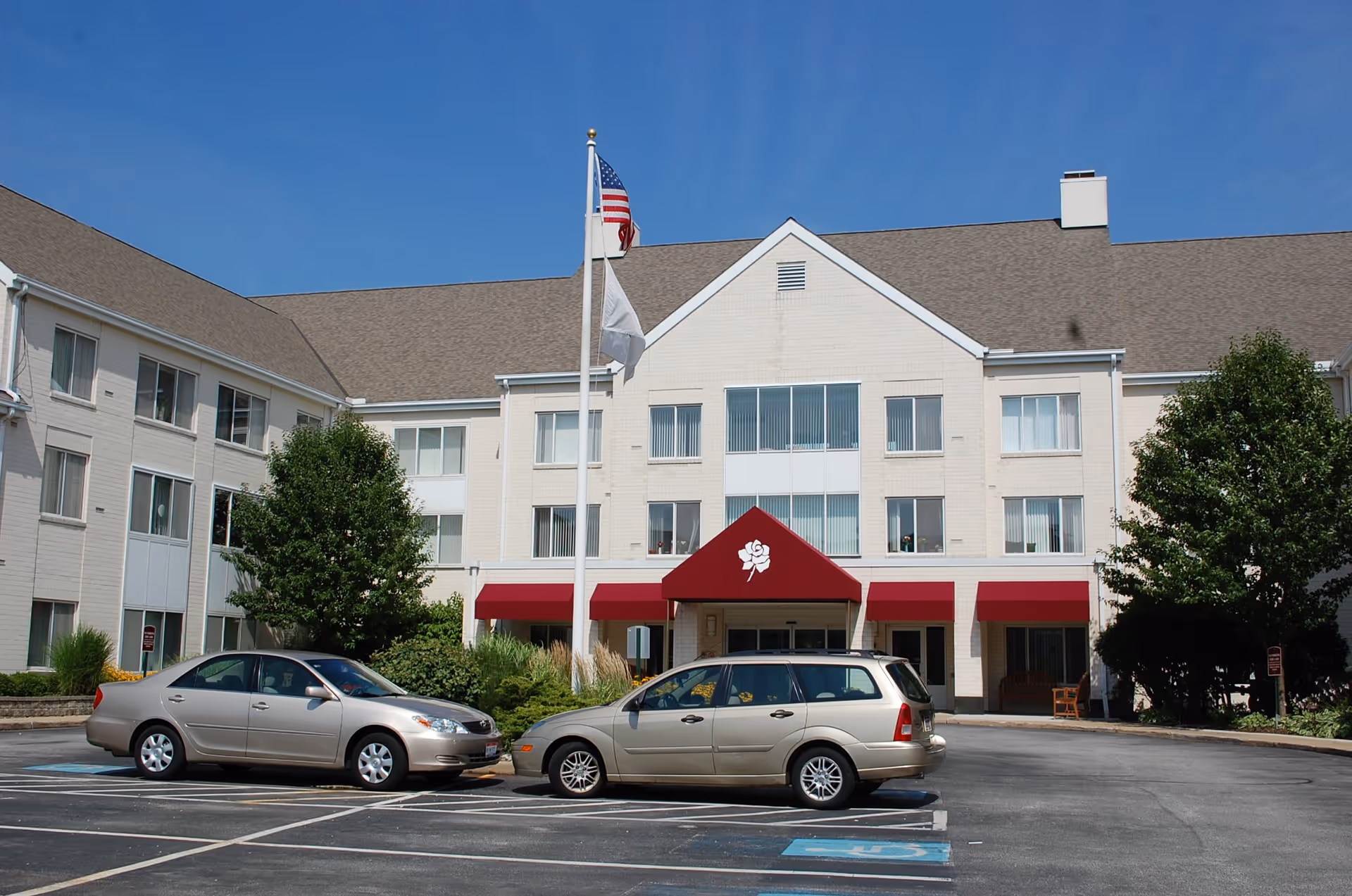 Front exterior view of a three-story senior living facility building with beige brick walls, multiple windows, and a maroon awning featuring a white rose logo above the entrance. Two cars are parked in front, and two flagpoles with an American flag and a white flag stand near the entrance. Trees and shrubs surround the building under a clear blue sky.