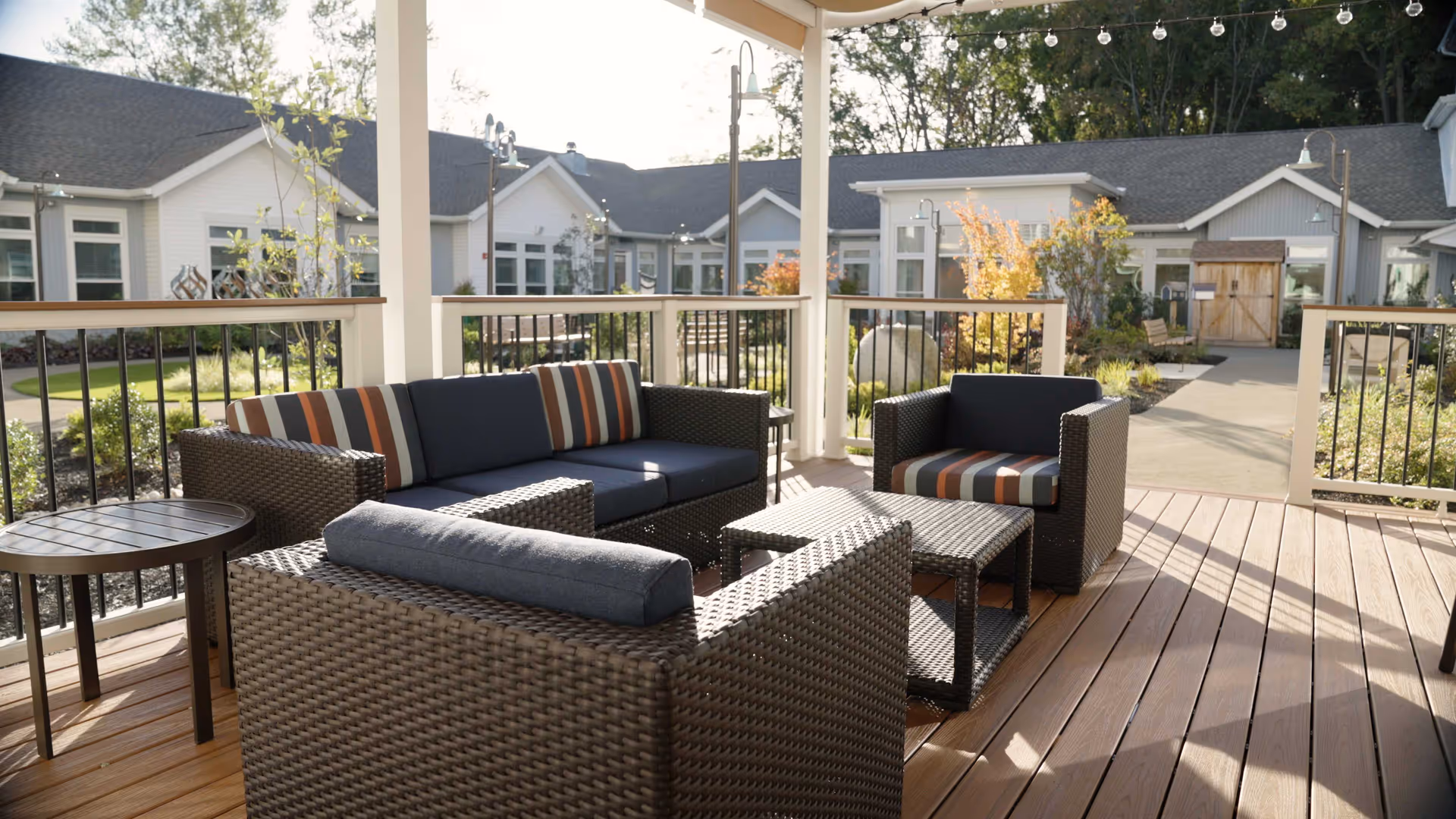 Covered outdoor patio with wicker sofa and chairs with striped cushions overlooking a landscaped courtyard and single-story building.