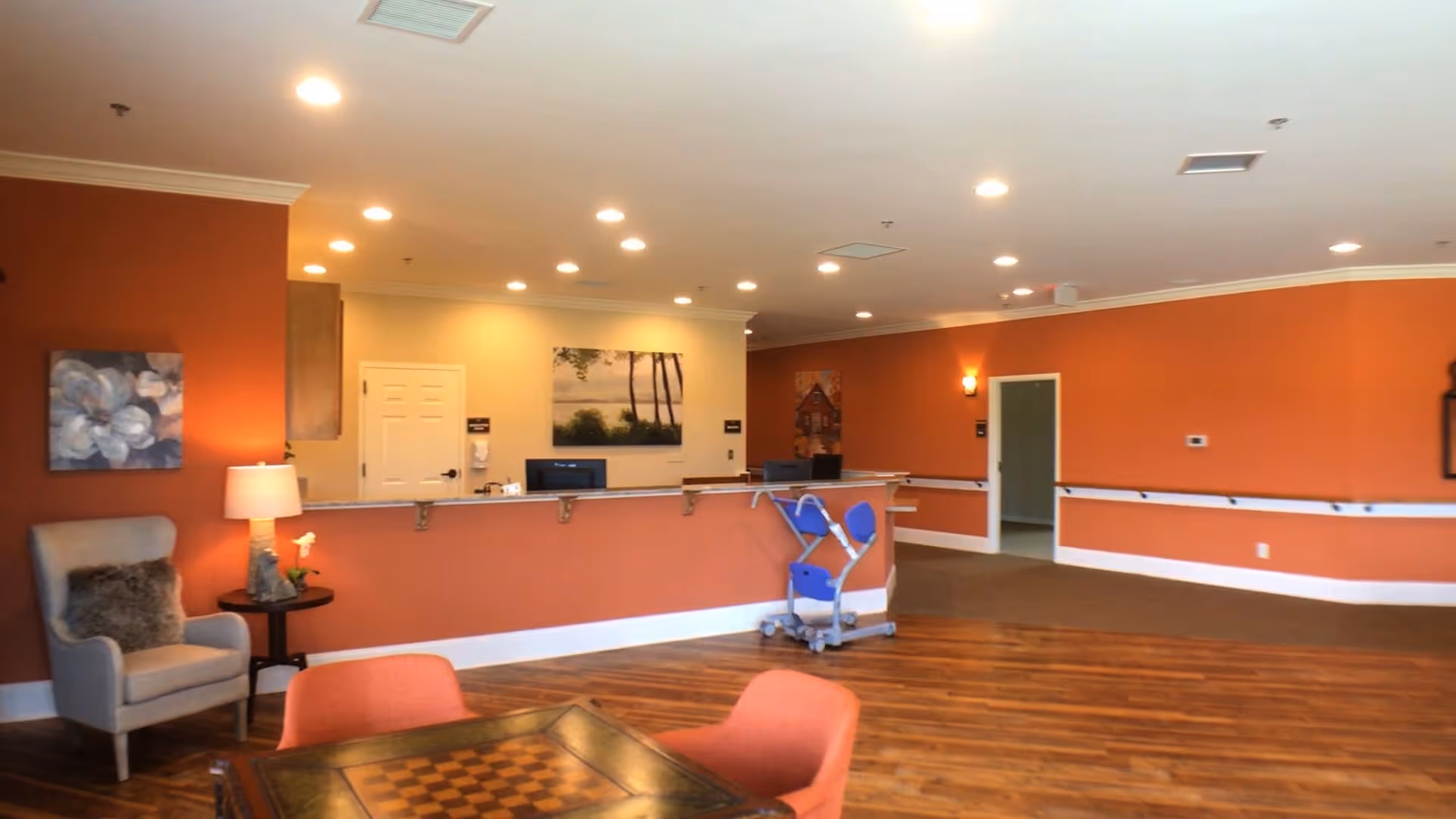 Interior view of a senior living facility reception area with orange walls, wooden flooring, a reception desk with two computer monitors, a blue mobility aid device, a gray armchair with a cushion, a side table with a lamp and flower, and wall art including paintings of flowers and trees.