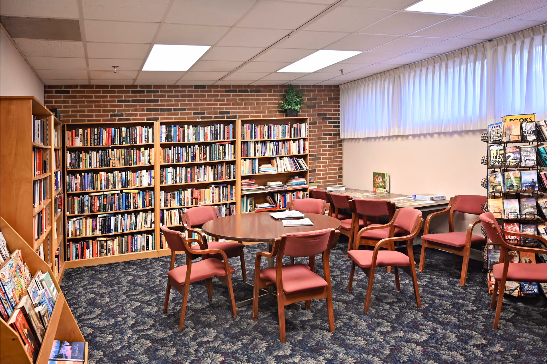 A cozy library room with wooden bookshelves filled with books along a brick wall. There is a round wooden table surrounded by eight red cushioned chairs in the center of the room. Additional chairs and tables are placed near a window with white curtains. A rotating rack with magazines and books is visible on the right side.