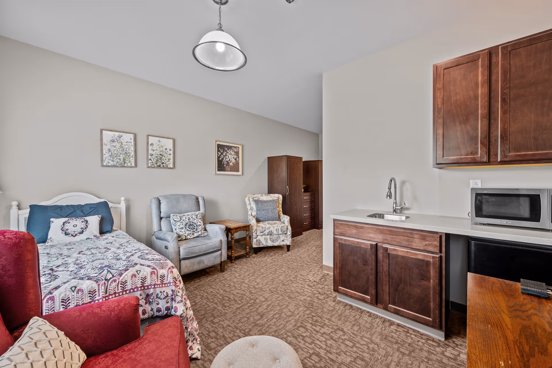 A senior living facility room featuring a single bed with patterned bedding, a red armchair, two additional armchairs with decorative pillows, a small wooden side table, and a kitchenette area with a sink, microwave, and dark wooden cabinets. The walls are light-colored with three framed floral artworks hanging above the bed.
