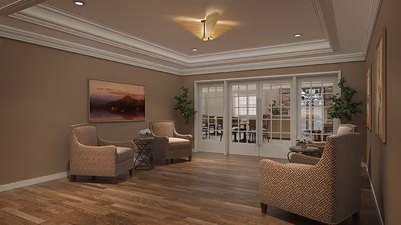 A cozy sitting area with three patterned armchairs arranged around two small round tables with flower arrangements. The room has wooden flooring, beige walls, and a ceiling with recessed lighting and a ceiling fan. French doors with glass panes lead to another room with more seating and plants.