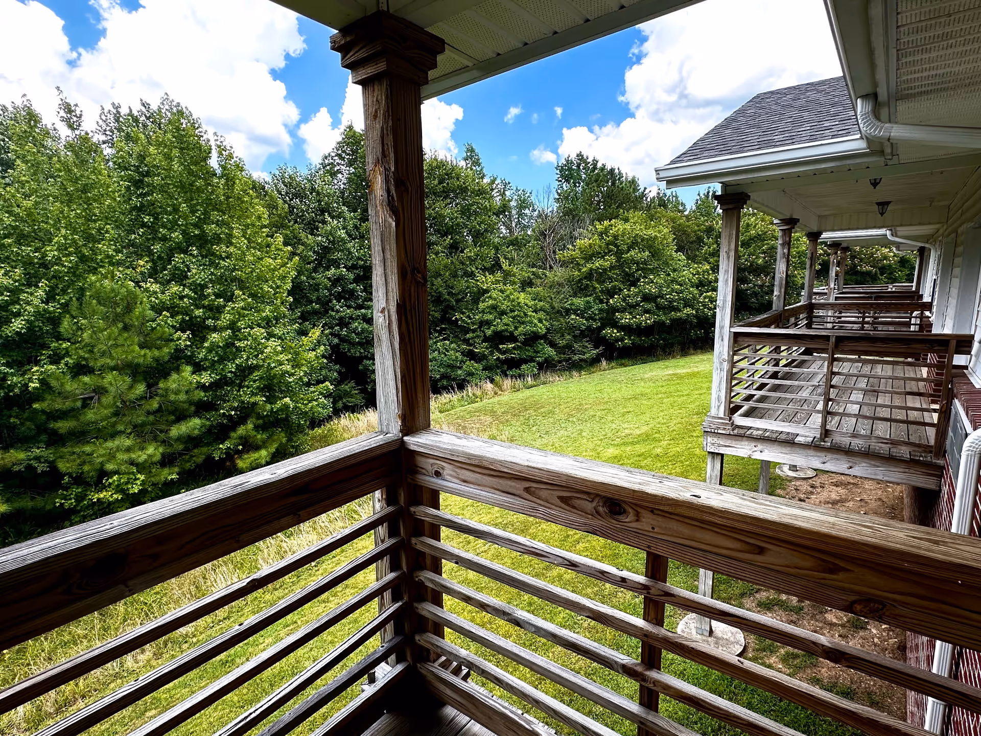 View from a wooden balcony overlooking a grassy lawn and dense green trees under a partly cloudy blue sky. Several similar balconies are attached to the side of a building with a sloped roof.