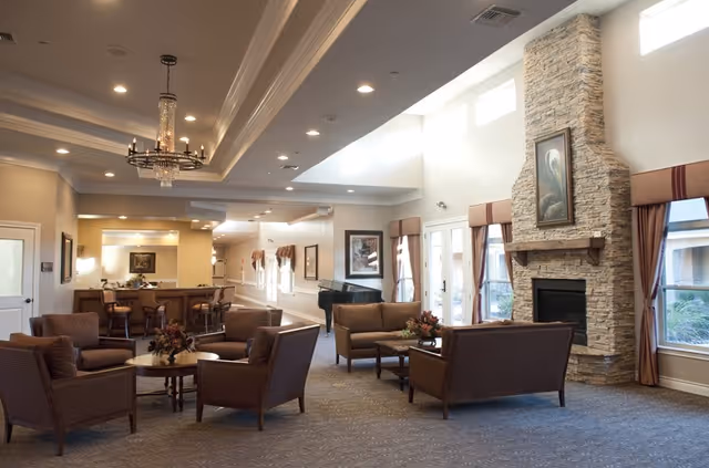 A spacious and well-lit living room area in an assisted living community featuring multiple brown upholstered chairs and sofas arranged around wooden coffee tables. The room has a high ceiling with recessed lighting and a chandelier, large windows with curtains, a stone fireplace with a painting above it, and a grand piano near the back. There is also a bar area with stools in the background.