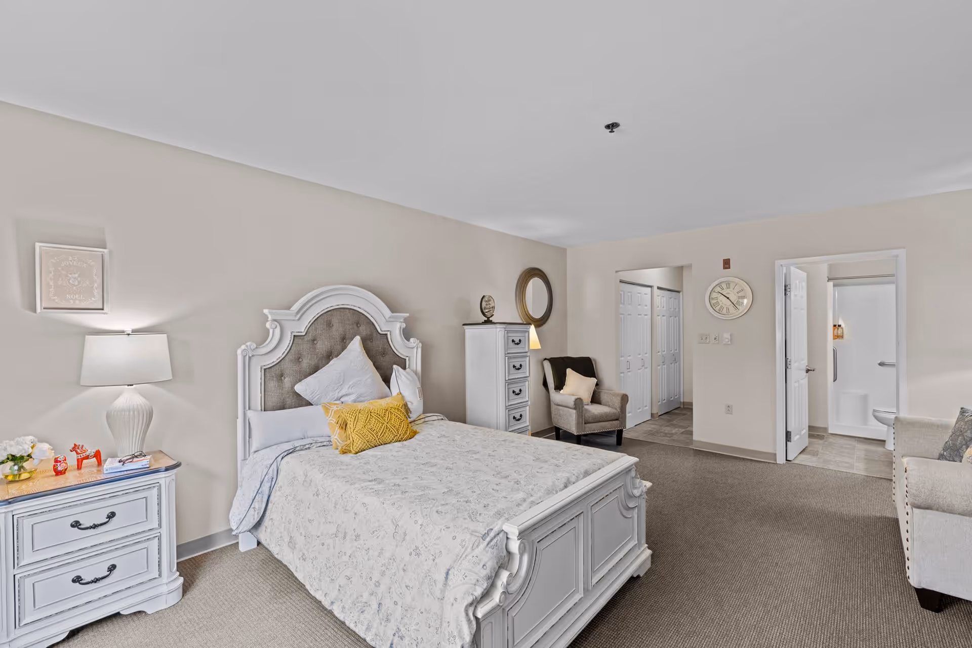 A bright and spacious senior living bedroom with a white ornate bed featuring a tufted headboard, a nightstand with a lamp and decorative items, a tall dresser, an armchair with a pillow, a wall clock, and an open door leading to a bathroom with a shower and toilet.