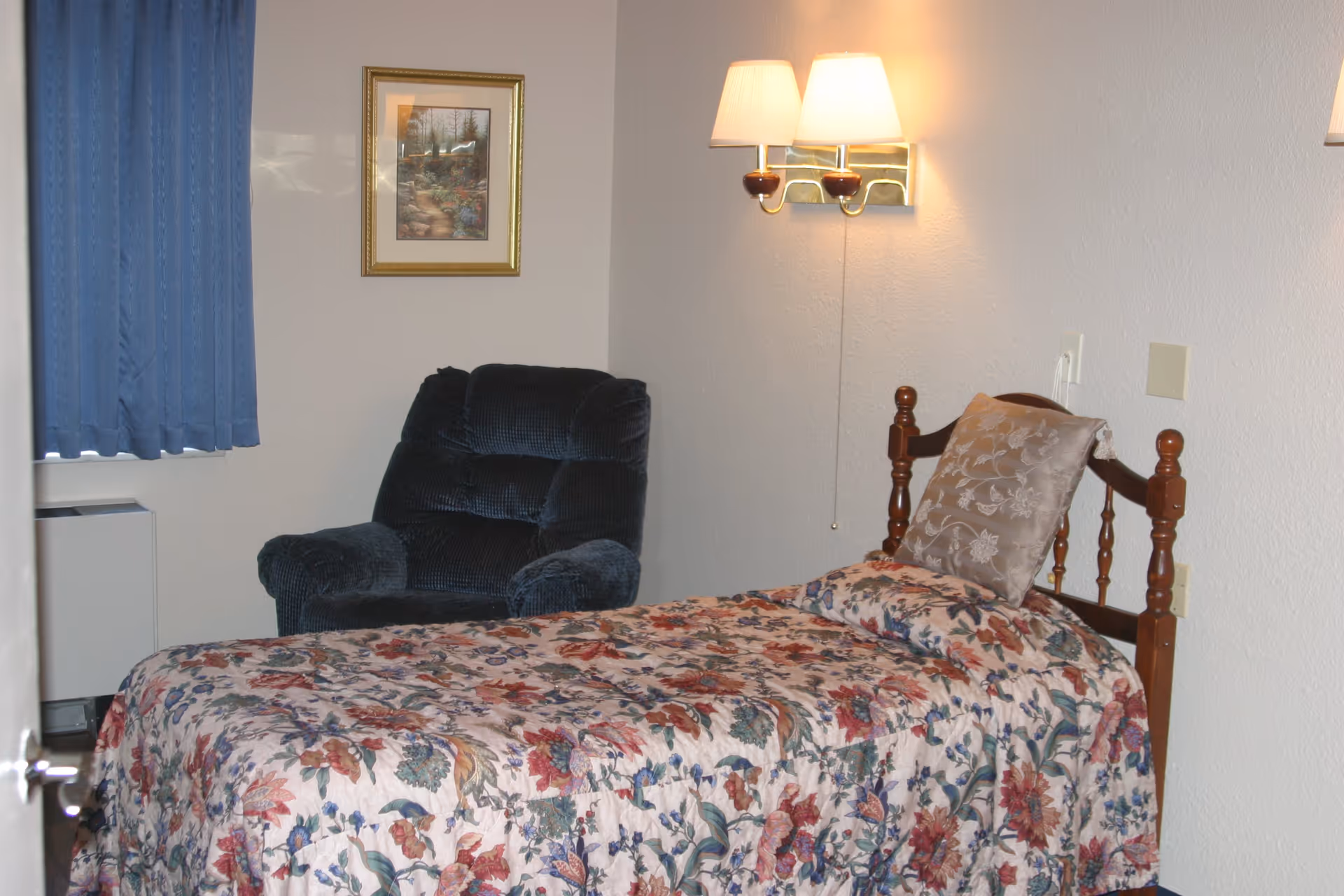 A small bedroom with a single wooden bed covered with a floral bedspread and a decorative pillow. Next to the bed is a dark blue upholstered armchair. The wall behind the bed has a double wall lamp with white lampshades and a framed picture of a garden path. Blue curtains cover the window on the left side of the room.
