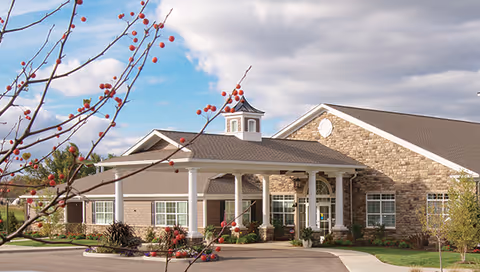 Exterior view of a single-story senior living facility building with stone and brick facade, white columns supporting a covered entrance, and a small cupola on the roof. There are trees and shrubs around the building and a partly cloudy sky in the background.