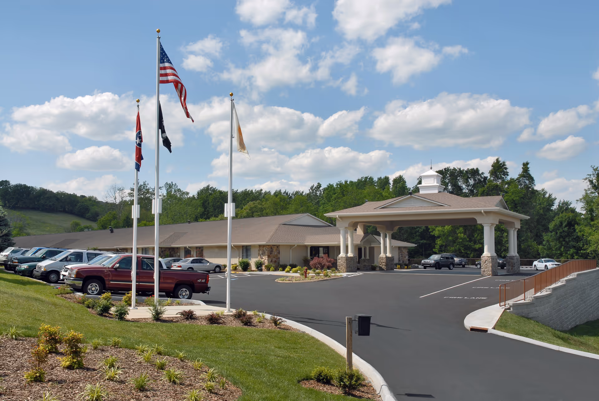 Exterior view of Life Care Center of Gray showing a single-story building with a covered entrance supported by columns. There are three flagpoles with flags in front of the building, a parking lot with several parked vehicles, and landscaped greenery surrounding the area under a partly cloudy sky.