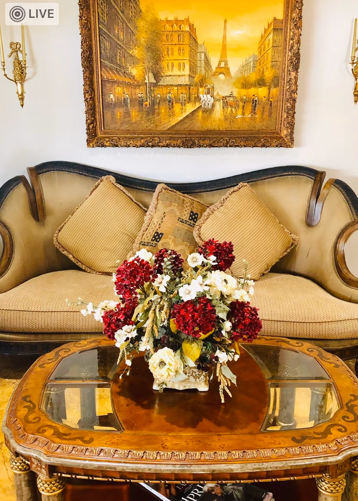 Ornate sofa with cushions and a floral centerpiece on a wooden coffee table beneath a framed Parisian painting.