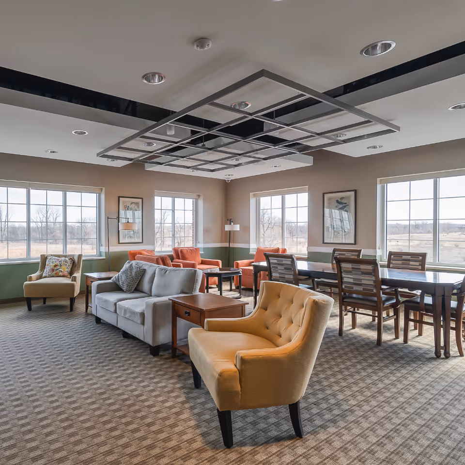 A spacious and well-lit common area in a senior living facility featuring multiple seating options including a beige armchair, a gray sofa with a patterned pillow, orange armchairs, and a long wooden dining table with chairs. Large windows allow natural light to fill the room, and framed artwork hangs on the walls. The ceiling has a modern grid design with recessed lighting.