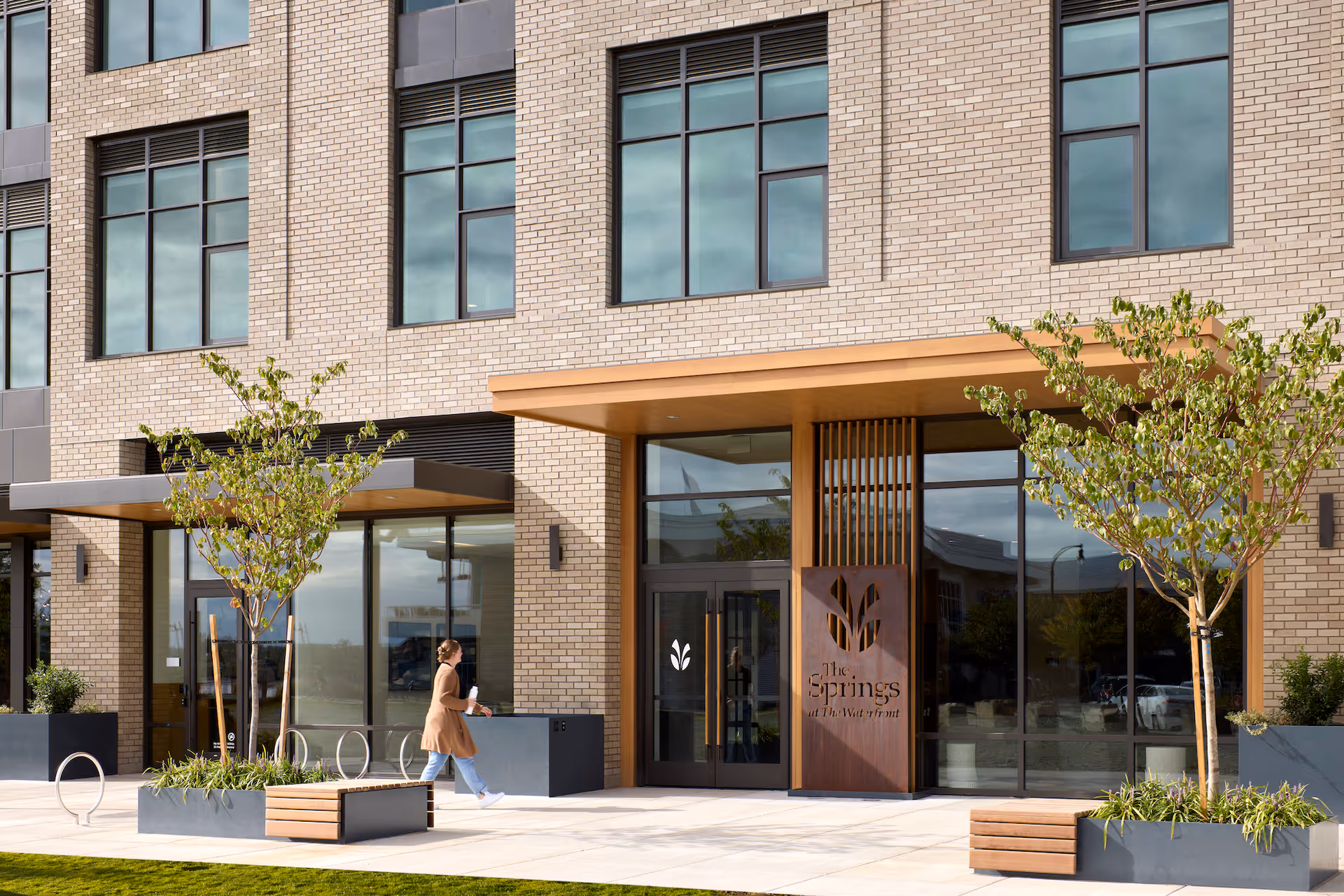 Exterior view of The Springs at The Waterfront building entrance with large windows, two small trees in planters, benches, and a person walking nearby.