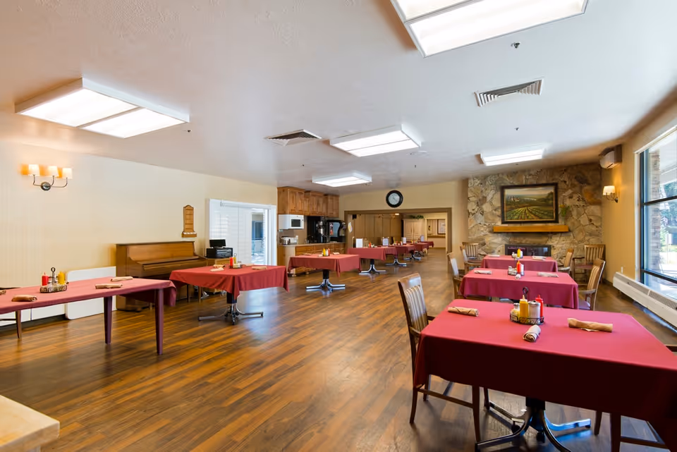 A spacious dining room with wooden floors and several tables covered with red tablecloths. Each table has rolled napkins and condiment holders. The room features a stone fireplace with a framed landscape painting above it, large windows letting in natural light, and a piano against one wall. The ceiling has recessed lighting panels.