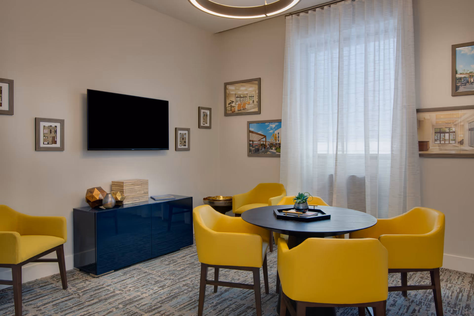 A cozy interior room with a round black table surrounded by five yellow armchairs. A small plant and a tray are on the table. The room has a large window with sheer white curtains, a flat-screen TV mounted on the wall, and several framed pictures hanging around it. A dark blue cabinet with decorative items sits below the TV.