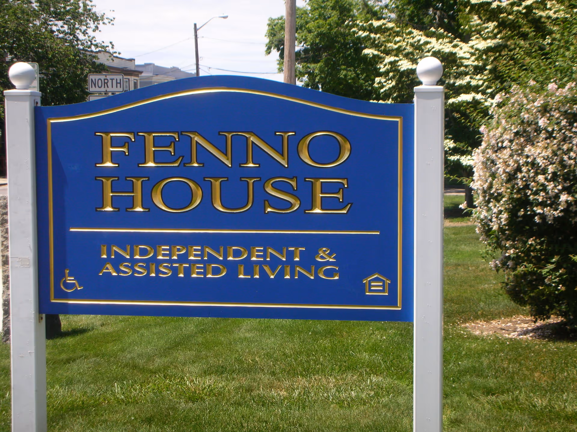 A blue and gold sign on a grassy area with white posts on each side. The sign reads 'Fenno House Independent & Assisted Living' with accessibility and housing icons. Trees and bushes are visible in the background.