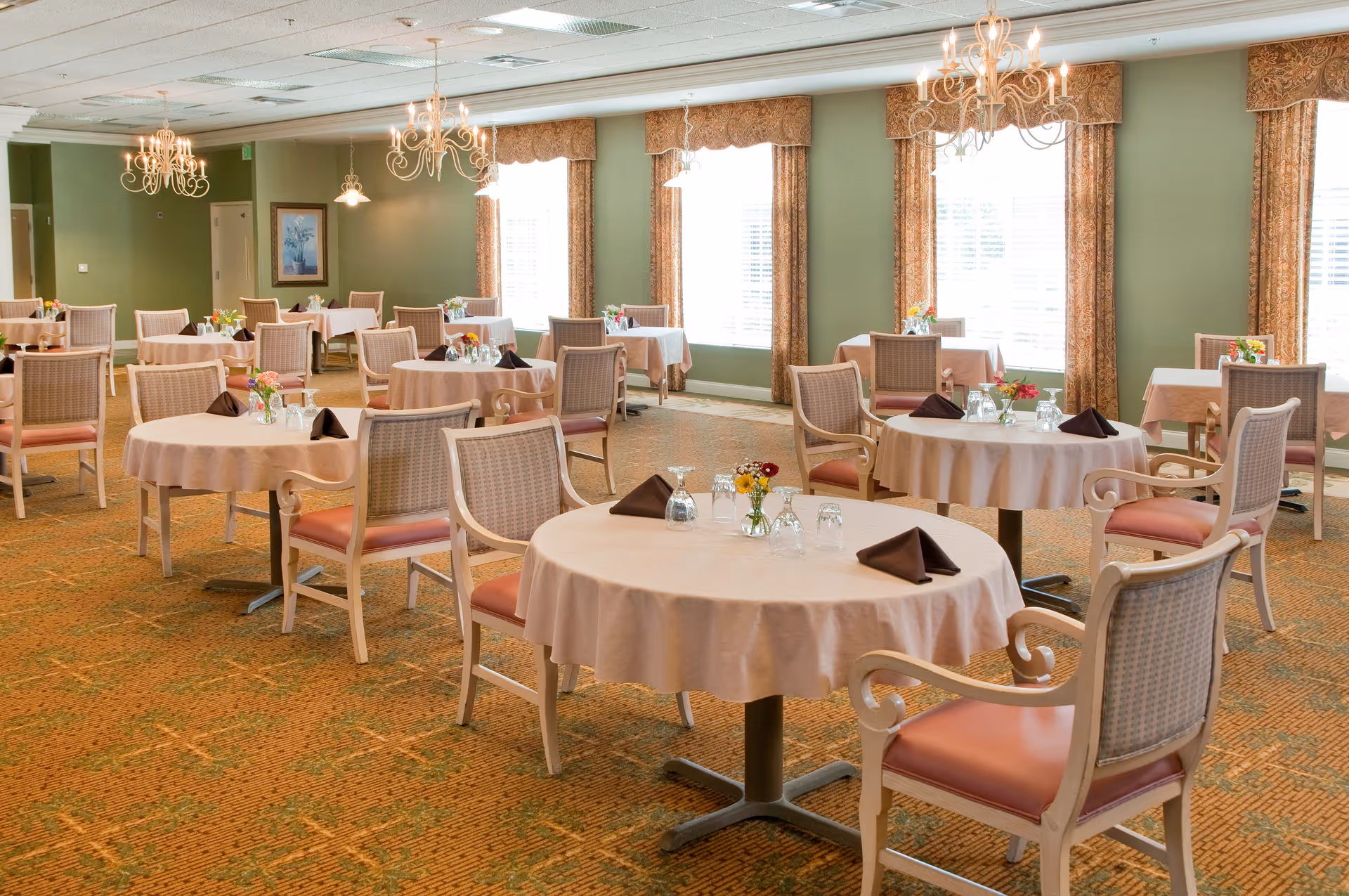 A bright dining room with multiple round tables covered with beige tablecloths, each set with brown folded napkins, glasses, and small flower arrangements. The room has green walls, large windows with patterned curtains, and elegant chandeliers hanging from the ceiling. The chairs have cushioned seats and backs with a light wood frame.