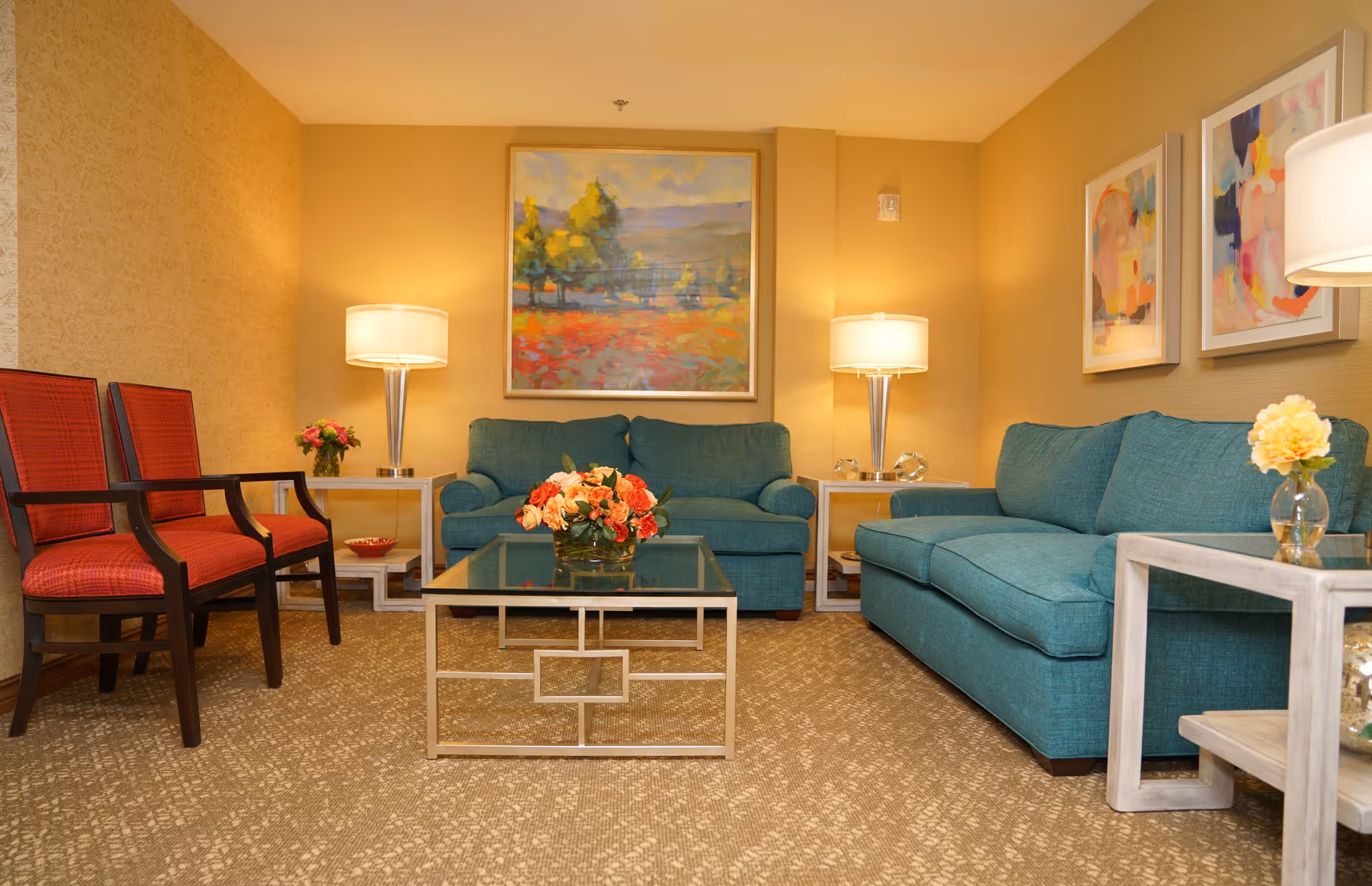 A cozy living room in a retirement community featuring two teal sofas, two red cushioned chairs, a glass coffee table with a floral centerpiece, two side tables with lamps, and colorful artwork on the walls.